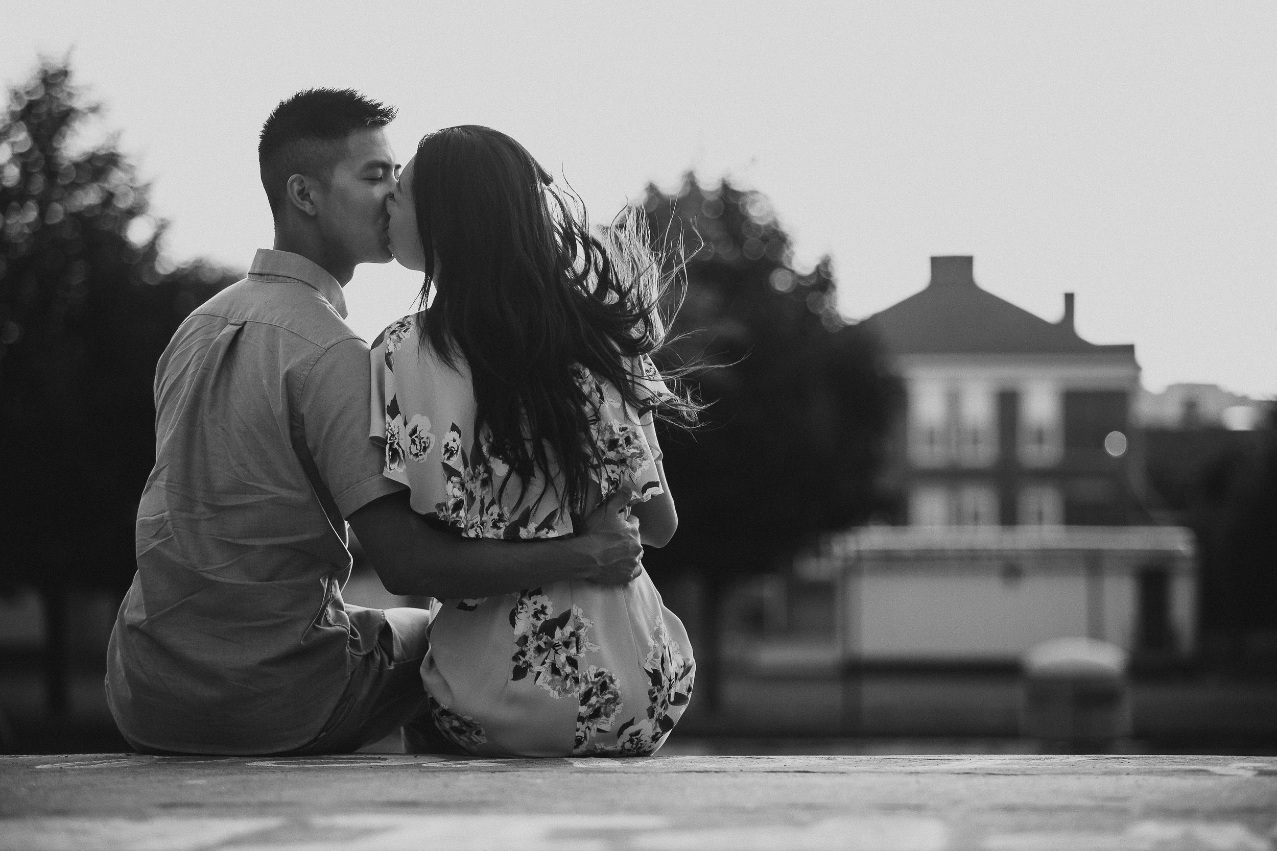 A romantic black and white photo series of a couple sitting together on a hill overlooking houses at sunset.