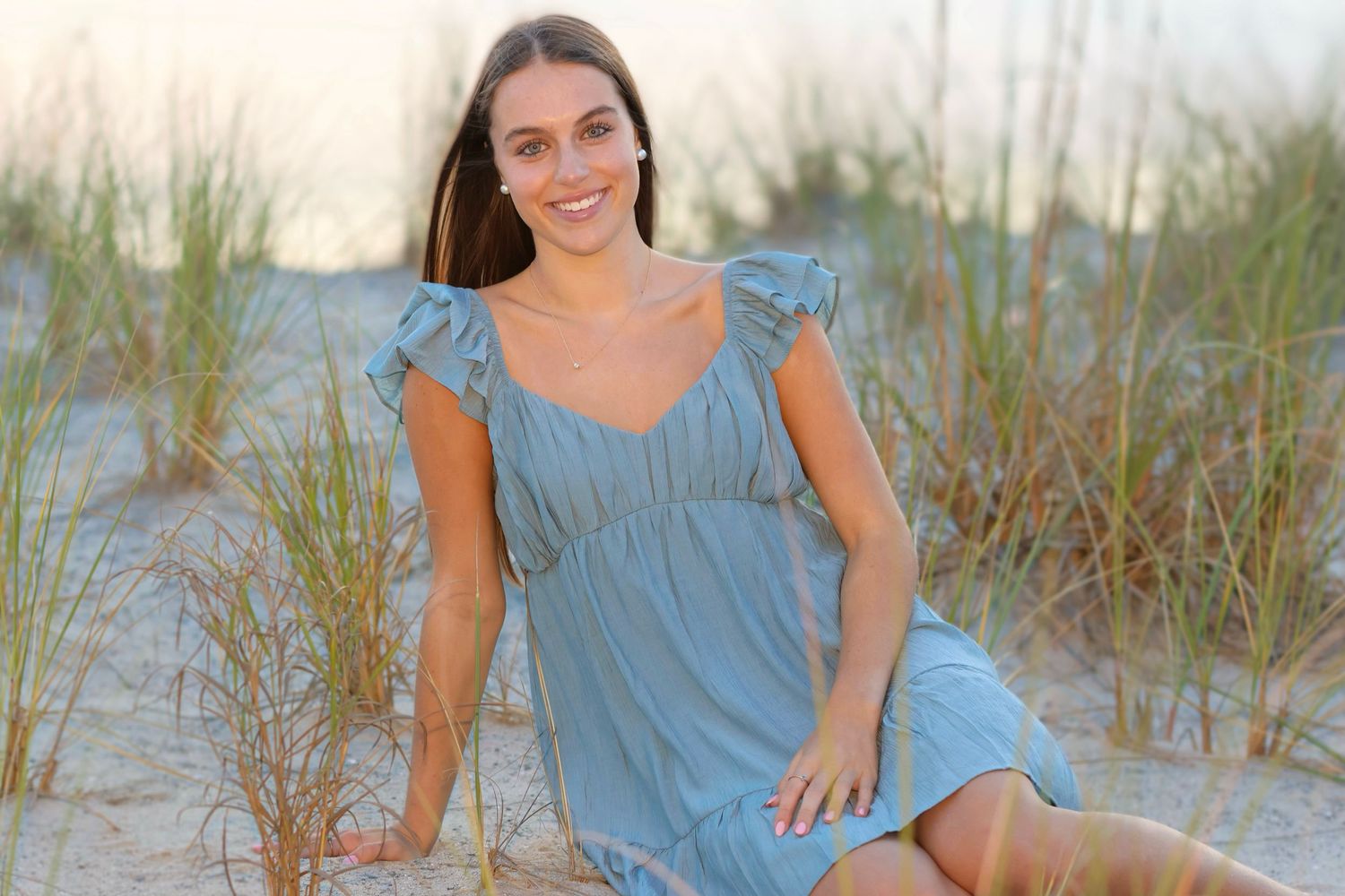 High school senior girl with short blue dress sitting in the sand with beach grass surrounding her at sunset.