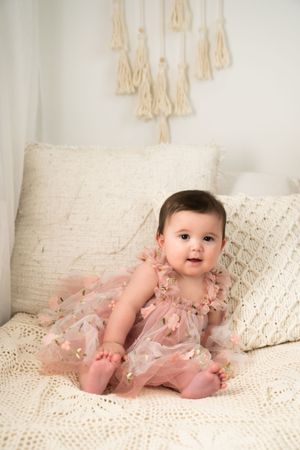 Baby in ruffled pink dress sits on cream couch with macrame wall hanging in background.