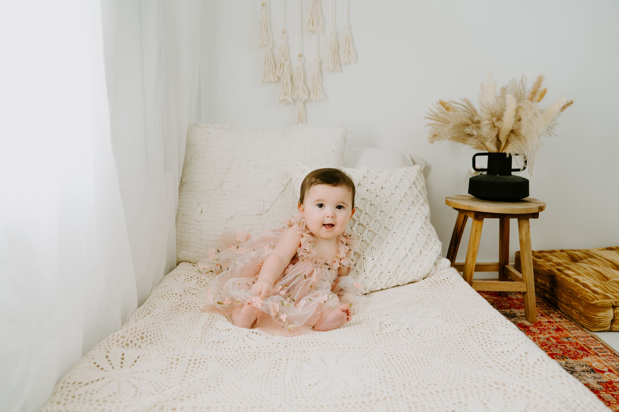 A baby in pink clothing sits on a white bed with macrame wall decor in the background.