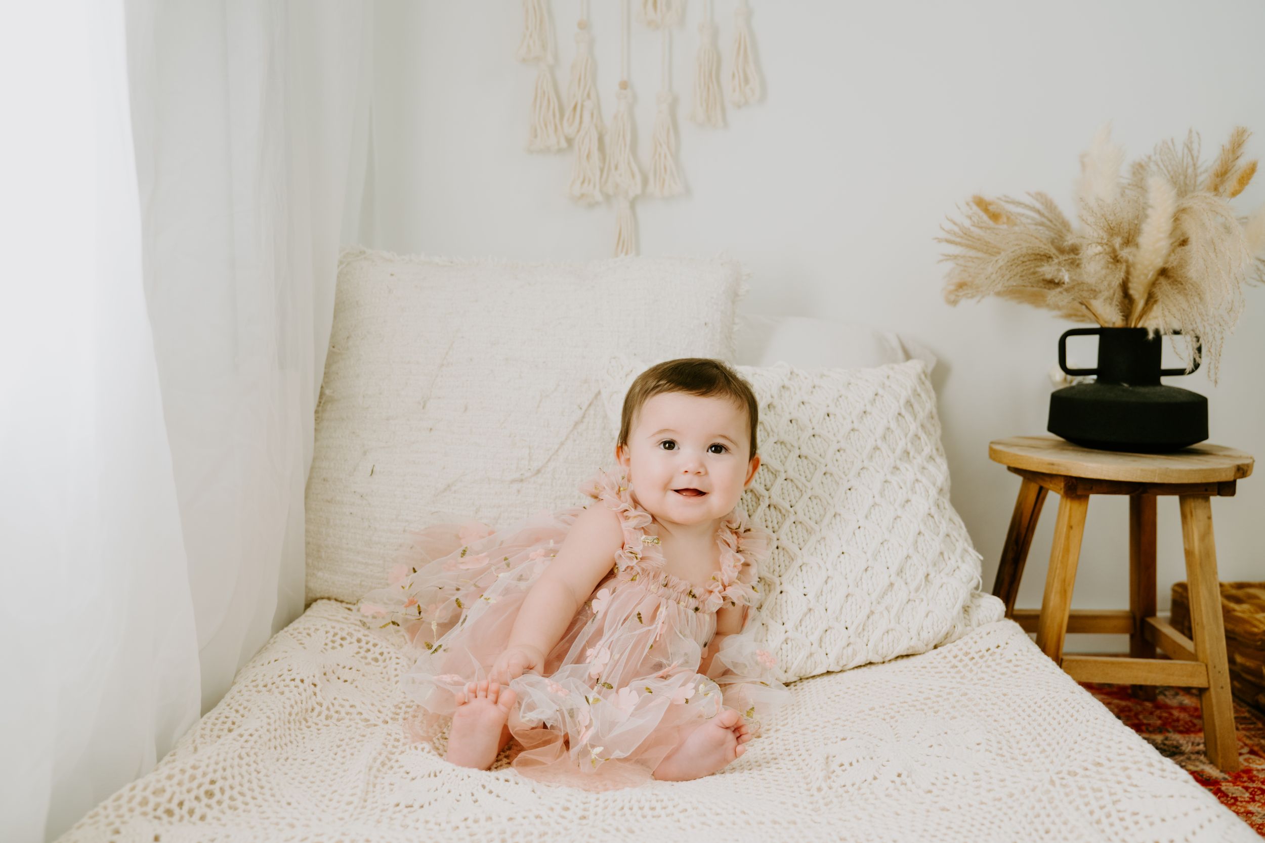 An infant wearing pink rests on cream colored bedding with boho decor visible.