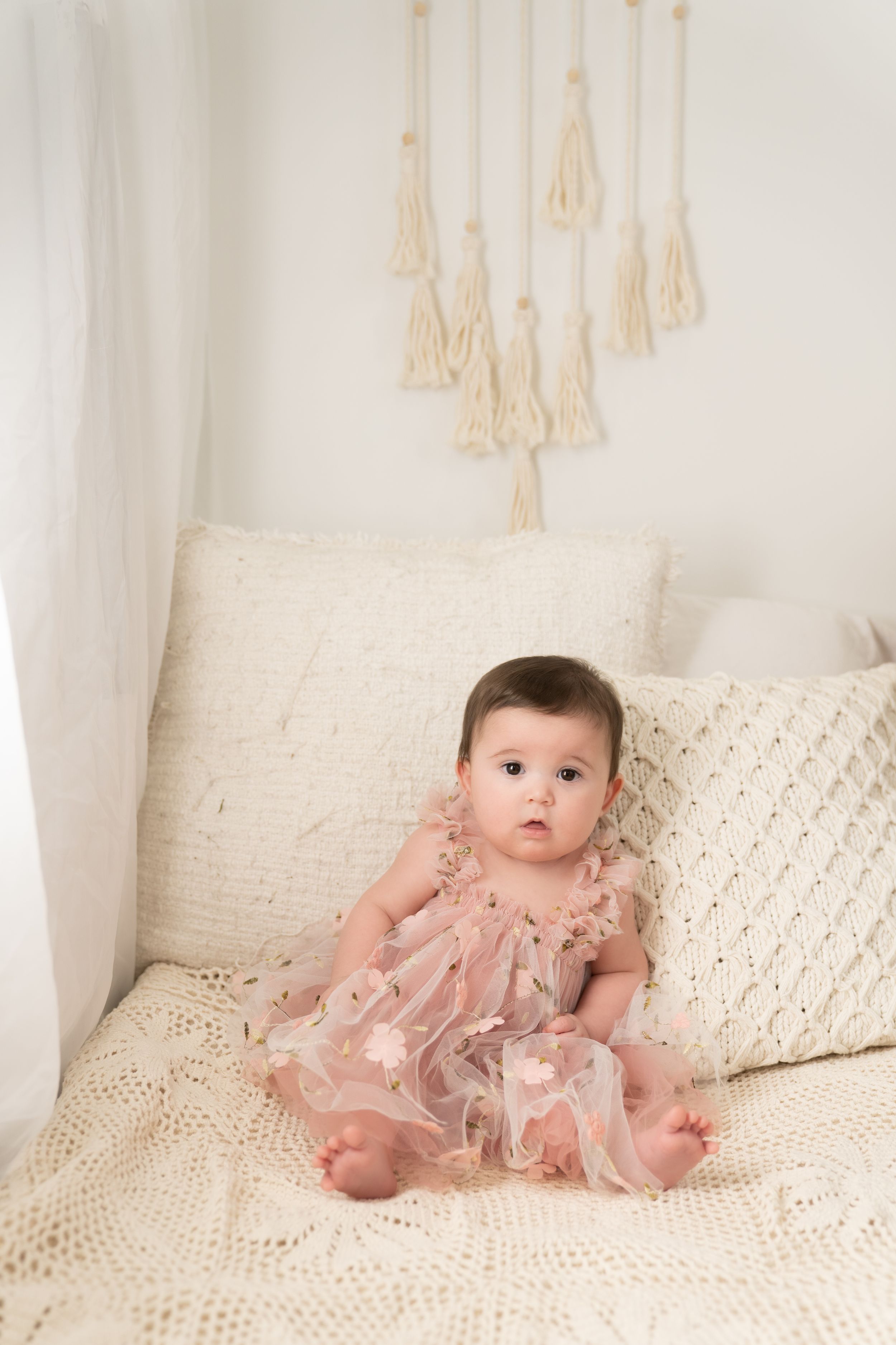 A baby sits upright on a cream colored bed wearing a pink tulle dress.