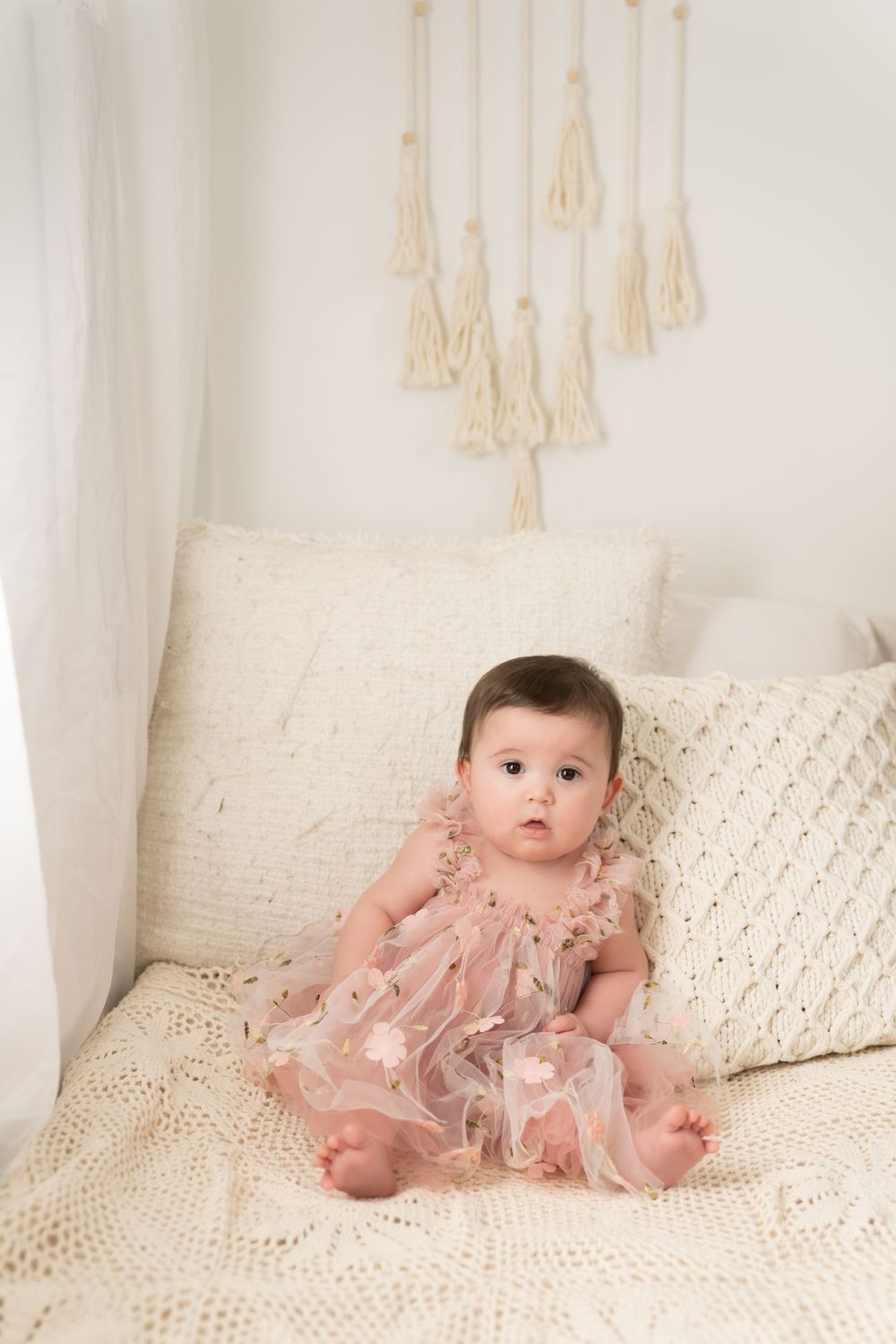 A baby sits upright on a cream colored bed wearing a pink tulle dress.