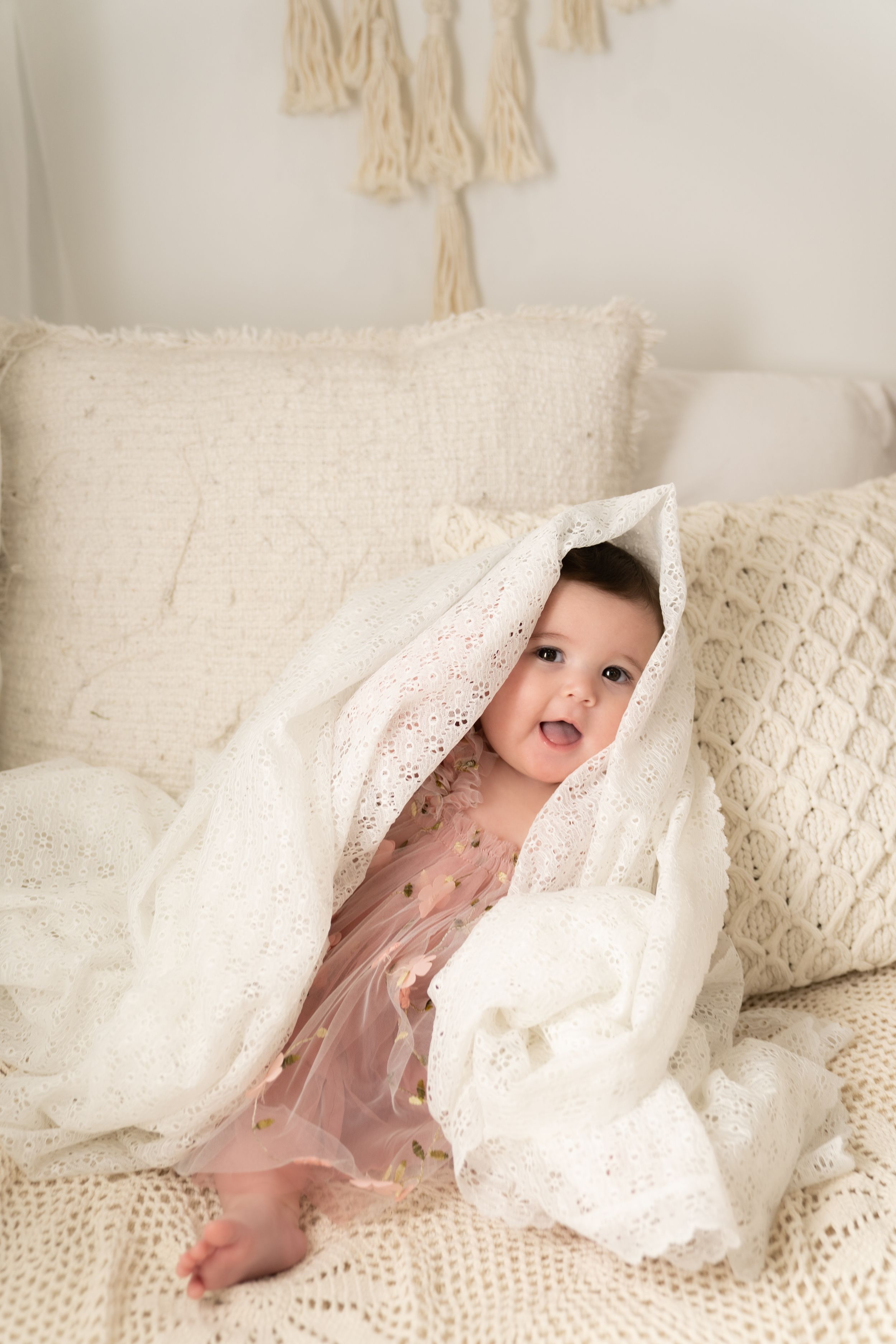 A laughing baby plays peek-a-boo with a white lace blanket on a cozy bed.