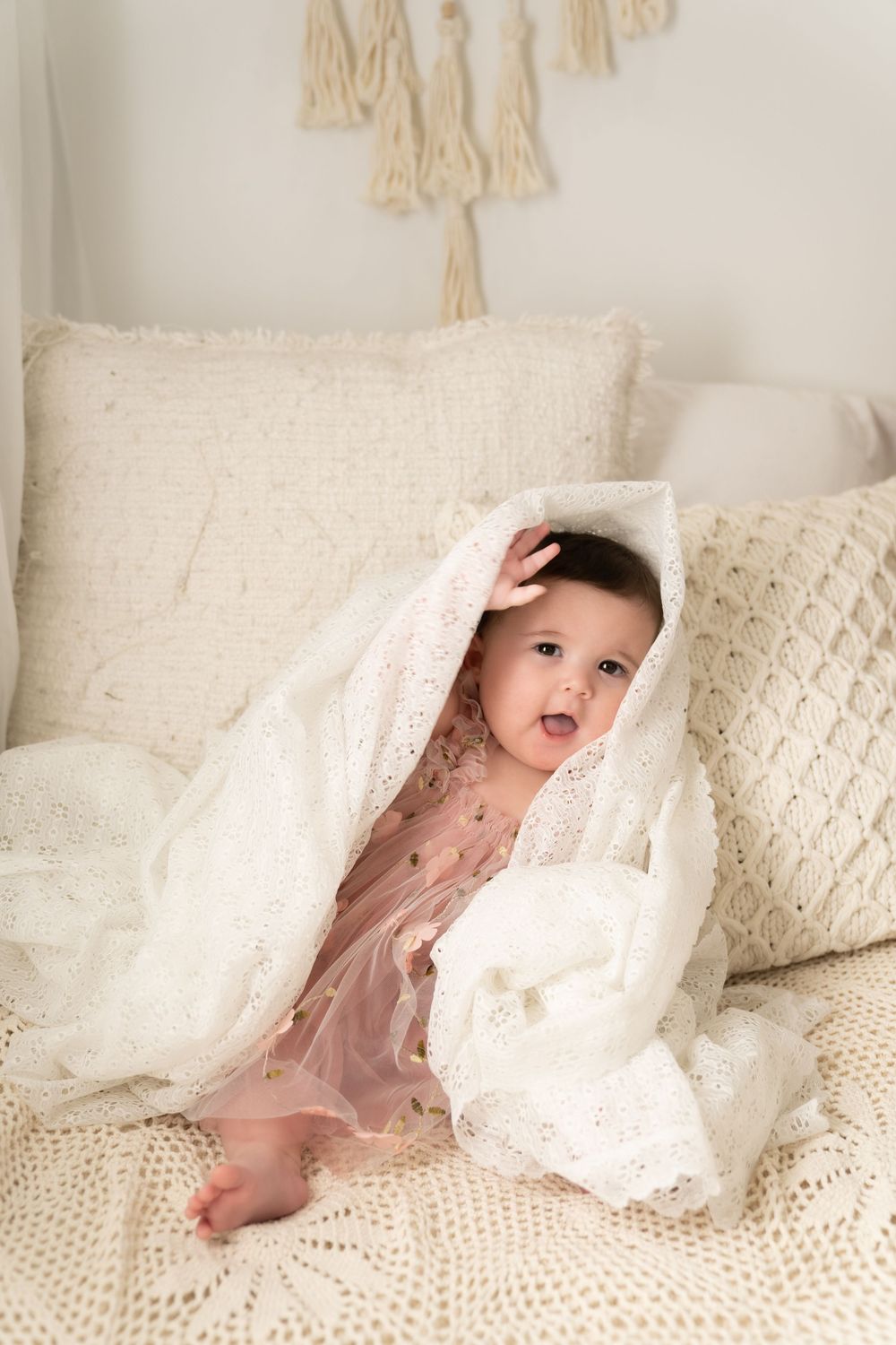 Baby in pink dress peeks out from under white lace blanket on couch with decorative pillows.