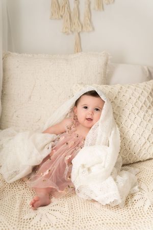 Baby wearing pink dress plays under white lace blanket on cream textured couch.