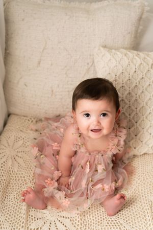 Baby in ruffled pink dress sits against cream pillows on textured blanket.
