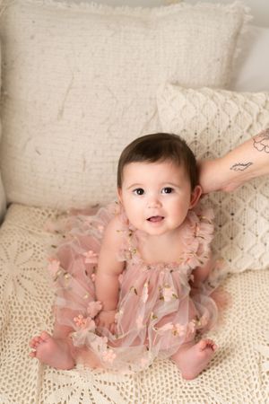 Baby in pink tulle dress sits smiling on cream knit blanket against neutral backdrop.