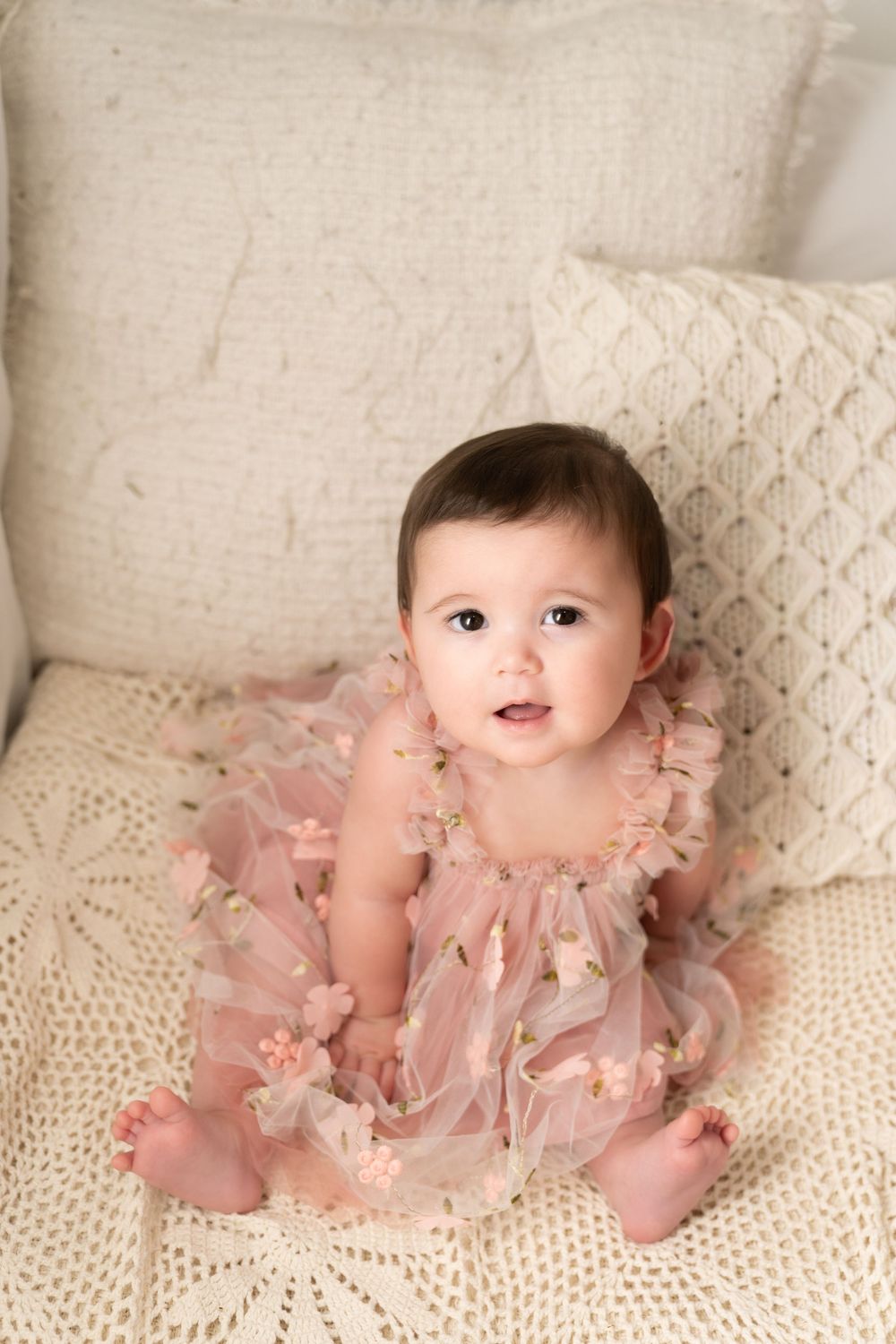 Baby wearing pink tulle dress poses on cream couch with decorative pillows.