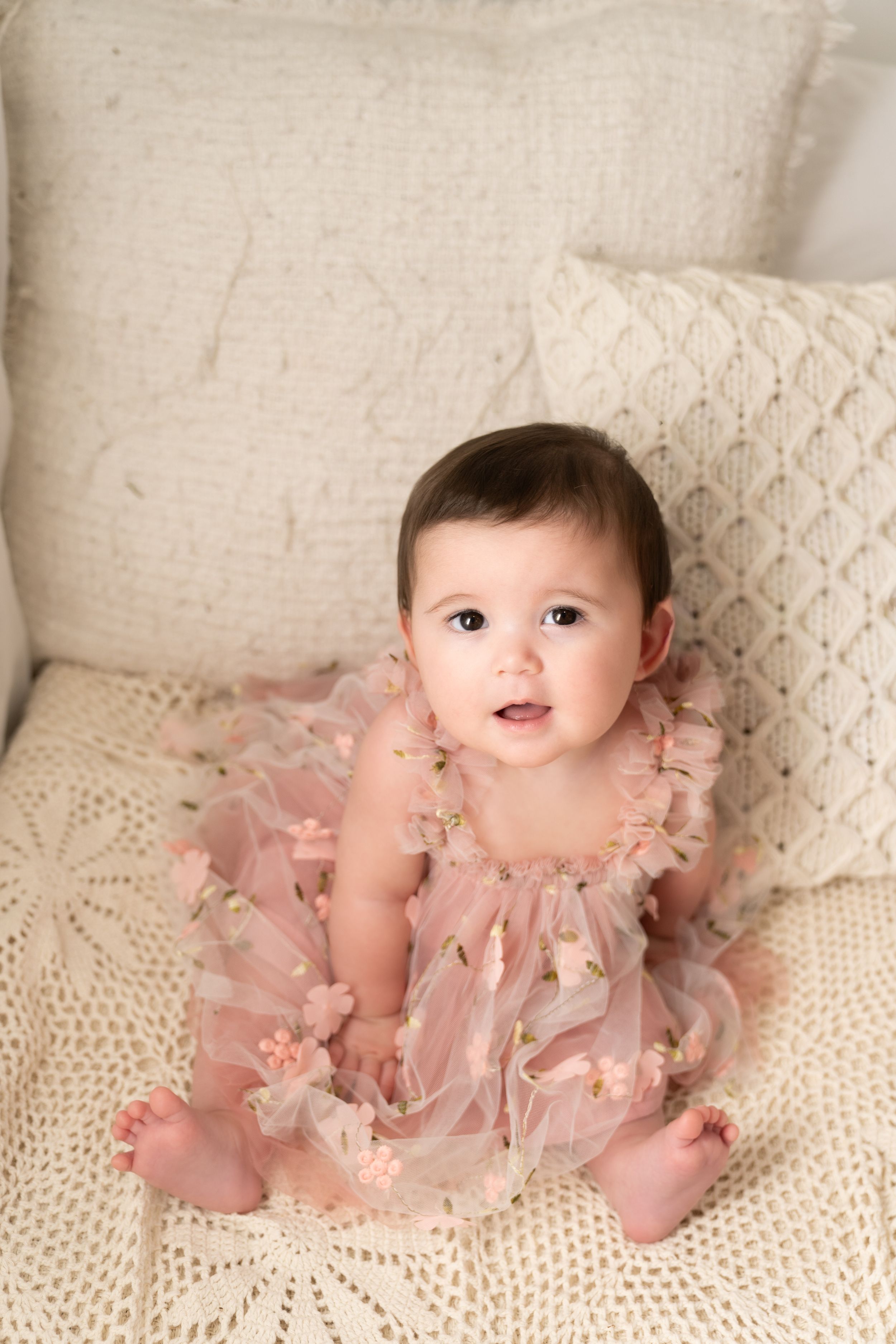 Baby wearing pink tulle dress poses on cream couch with decorative pillows.