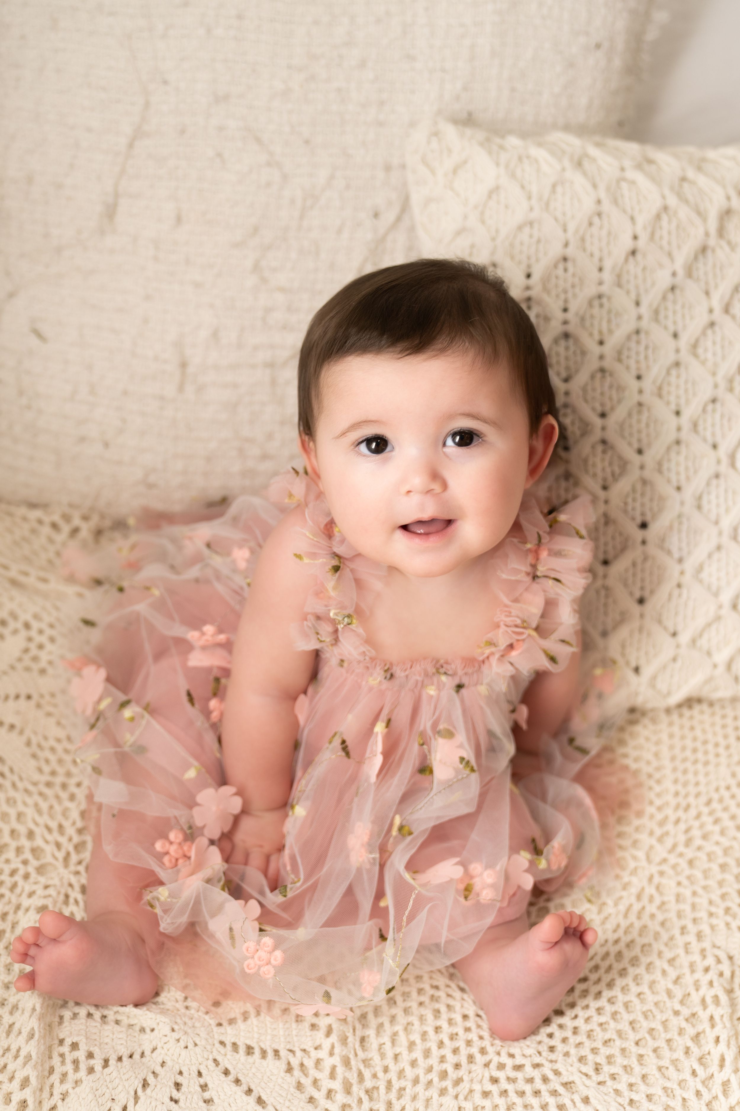 Baby wearing pink tulle dress sits smiling on cream knit blanket with decorative pillows.