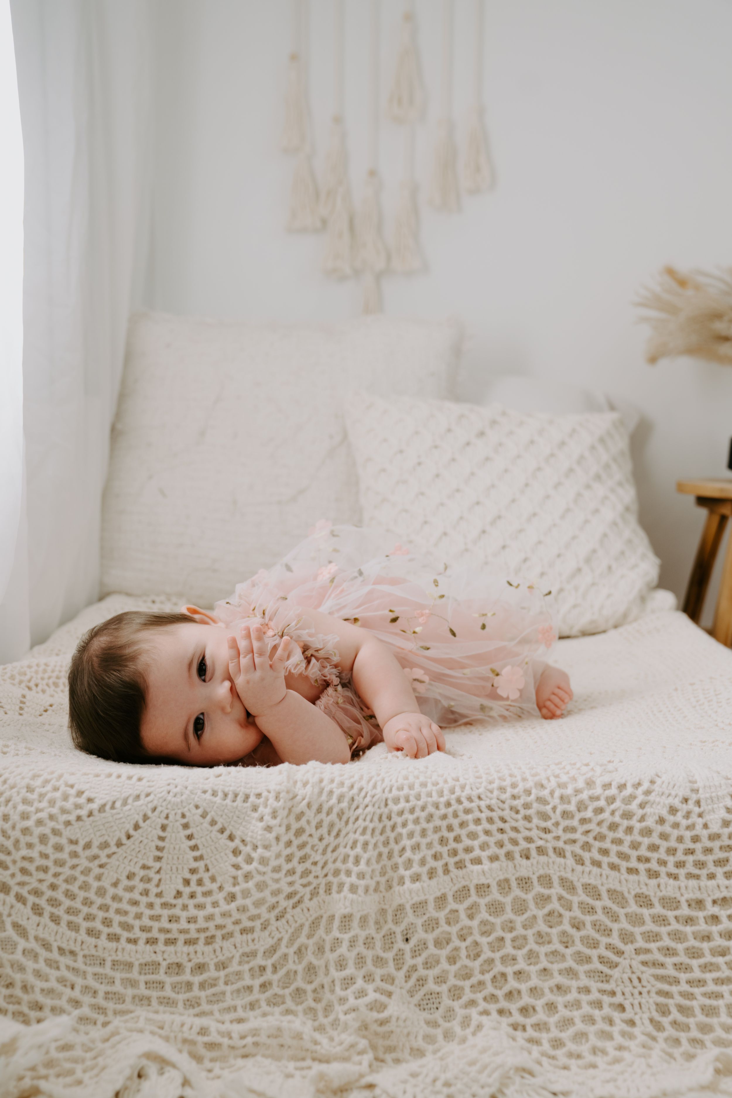 Baby in pink tulle dress lying on white textured blanket with macrame wall decor.