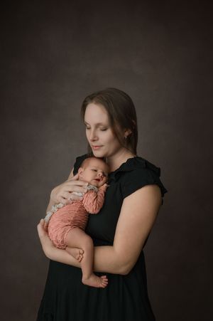 A woman in a black dress cradles and embraces a newborn baby wearing a soft peach-colored outfit in a dark studio setting.