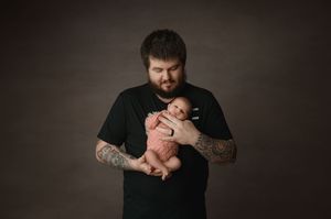 A bearded man in a black t-shirt with tattoos gently cradles a newborn baby in a minimalist portrait studio setting.