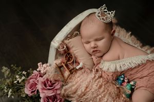 Sleeping newborn wearing crown accessory posed with pink roses and lace on soft beige backdrop.