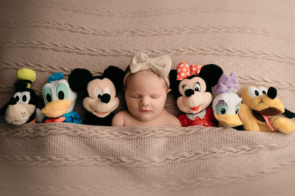 A baby sleeps peacefully surrounded by Disney character plush toys including Mickey Mouse, Minnie Mouse, and Goofy.