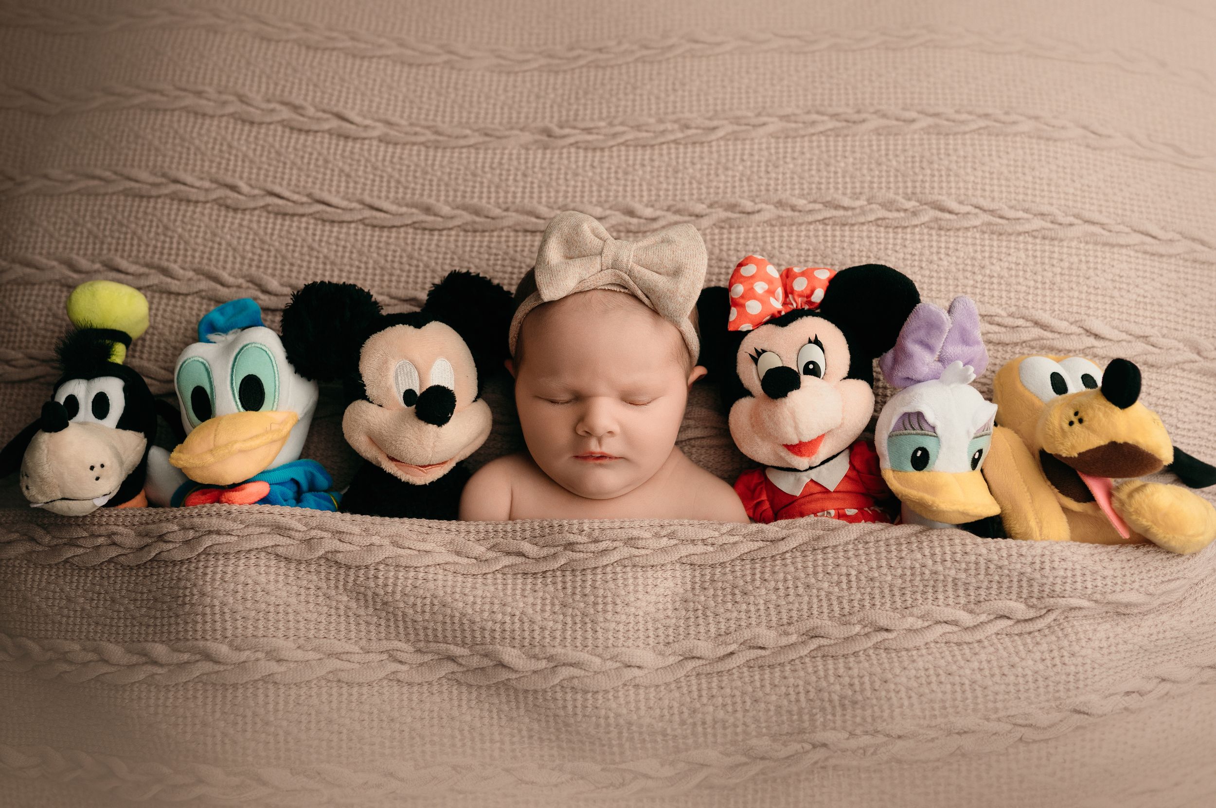 A baby sleeps peacefully surrounded by Disney character plush toys including Mickey Mouse, Minnie Mouse, and Goofy.