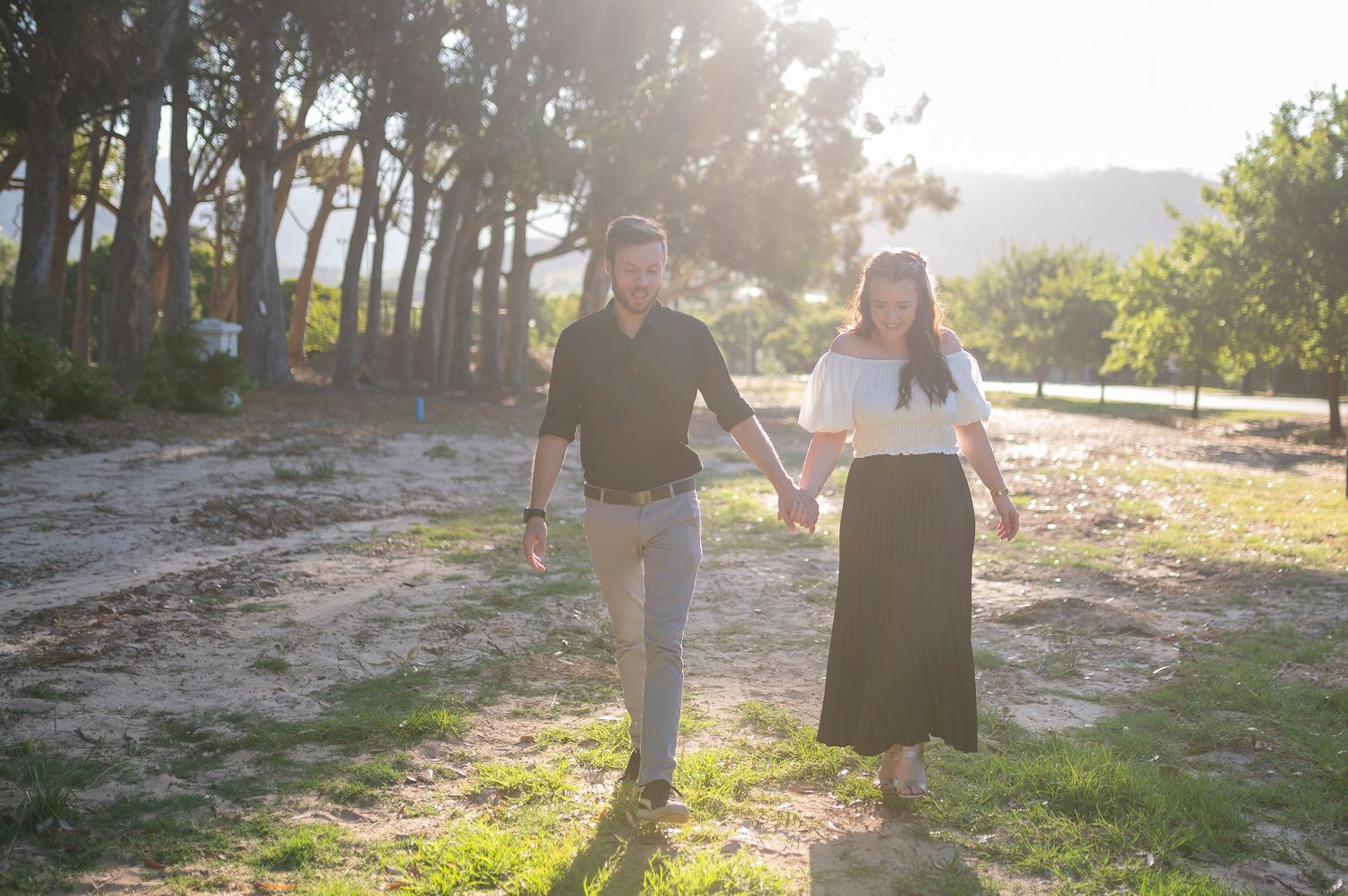 Couple walks hand in hand on a sunlit path through trees during golden hour at sunset.