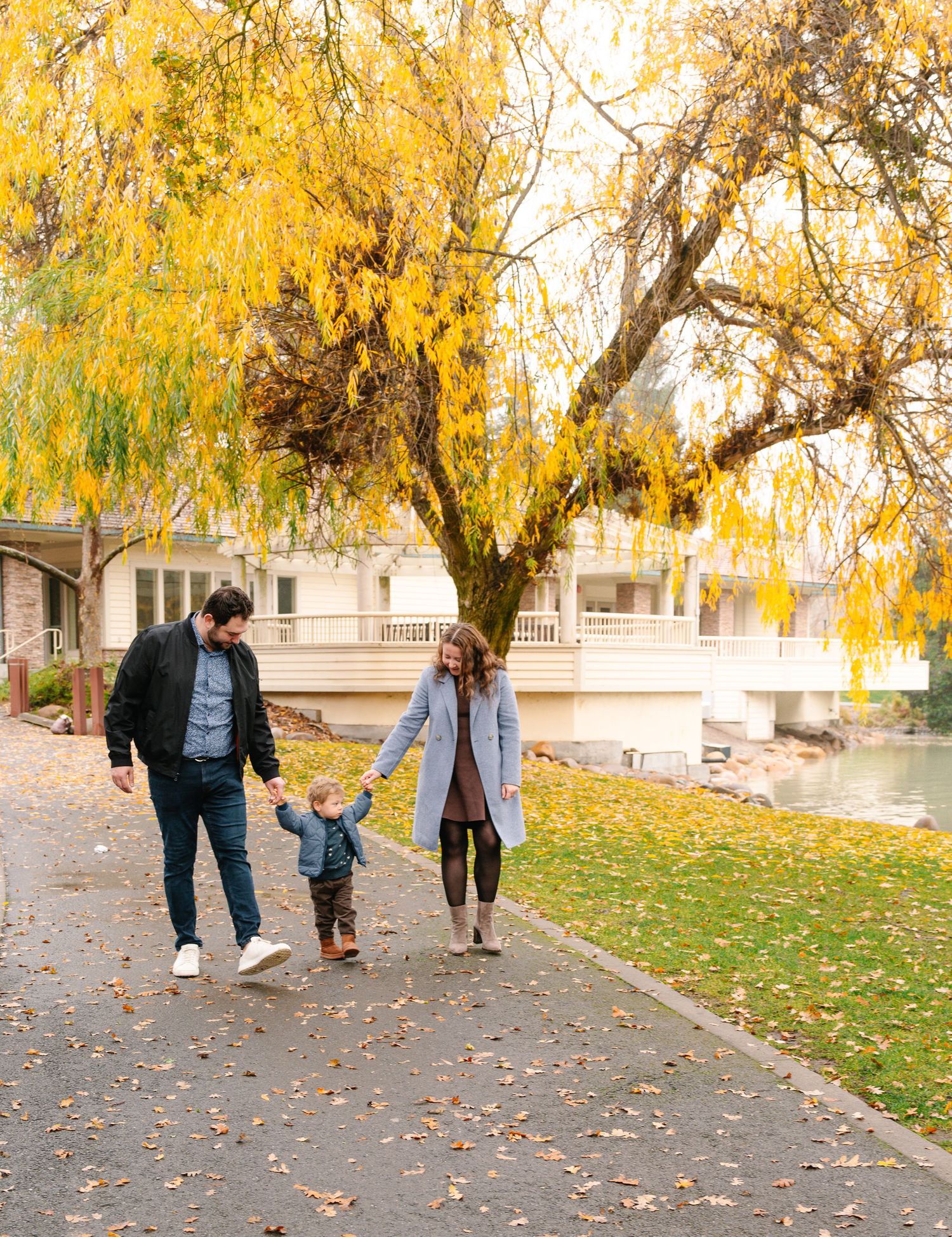 A family walks together under a golden autumn willow tree on a path by the water as fallen leaves scatter the ground.