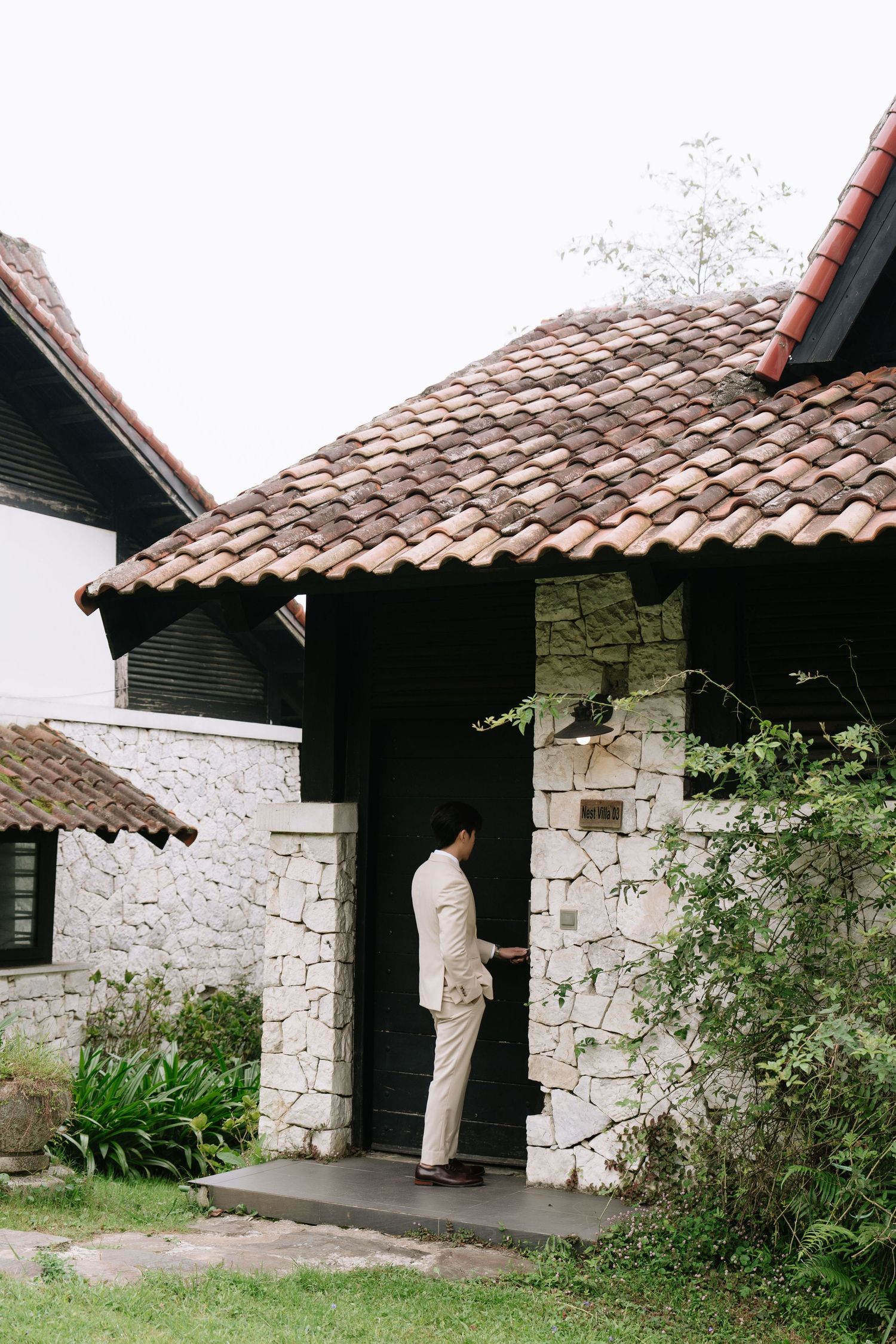 Groom in beige suit standing at entrance of stone villa with terracotta tile roof surrounded by greenery.