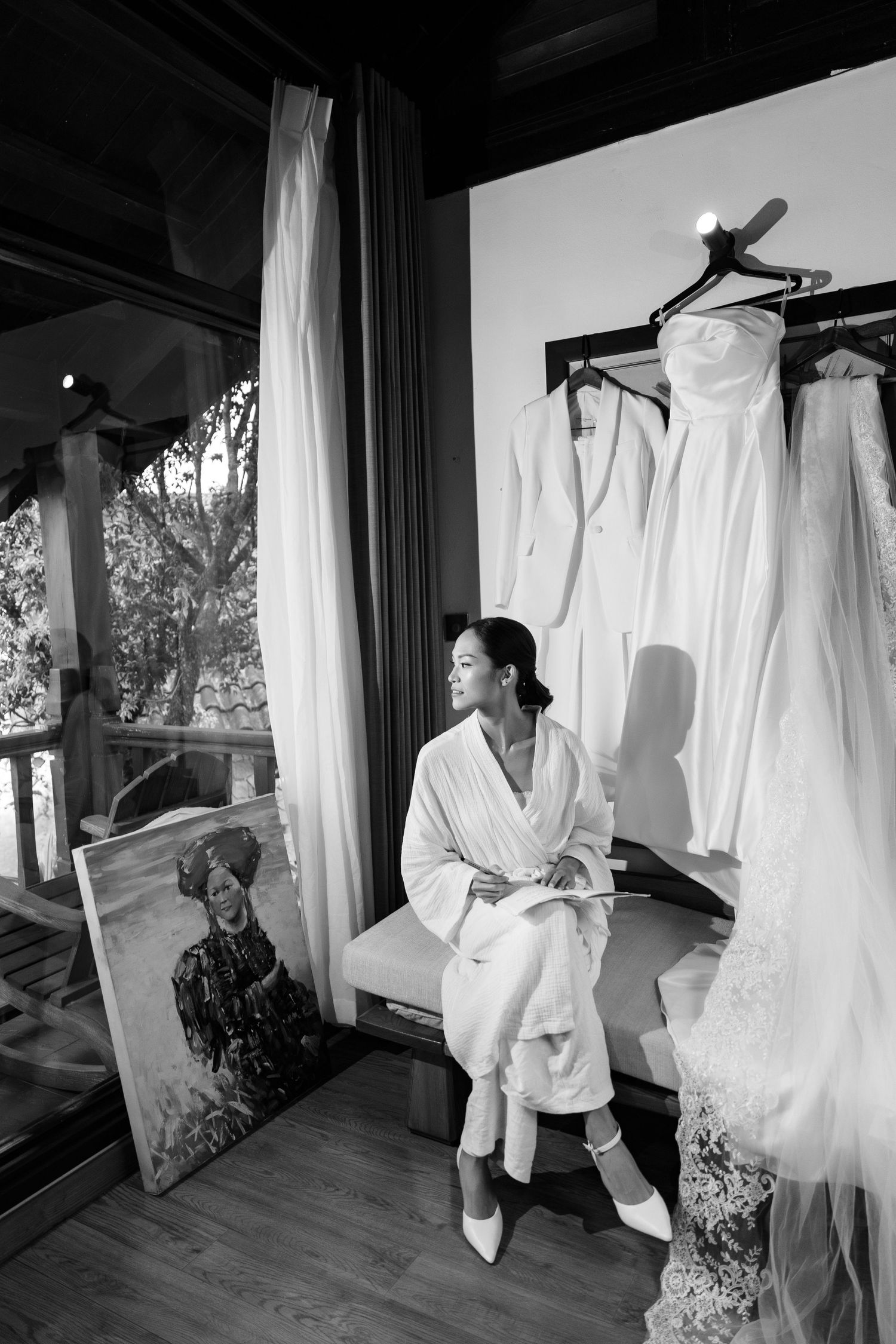 Bride in white robe sitting contemplatively beside hanging wedding dresses in black and white photo.
