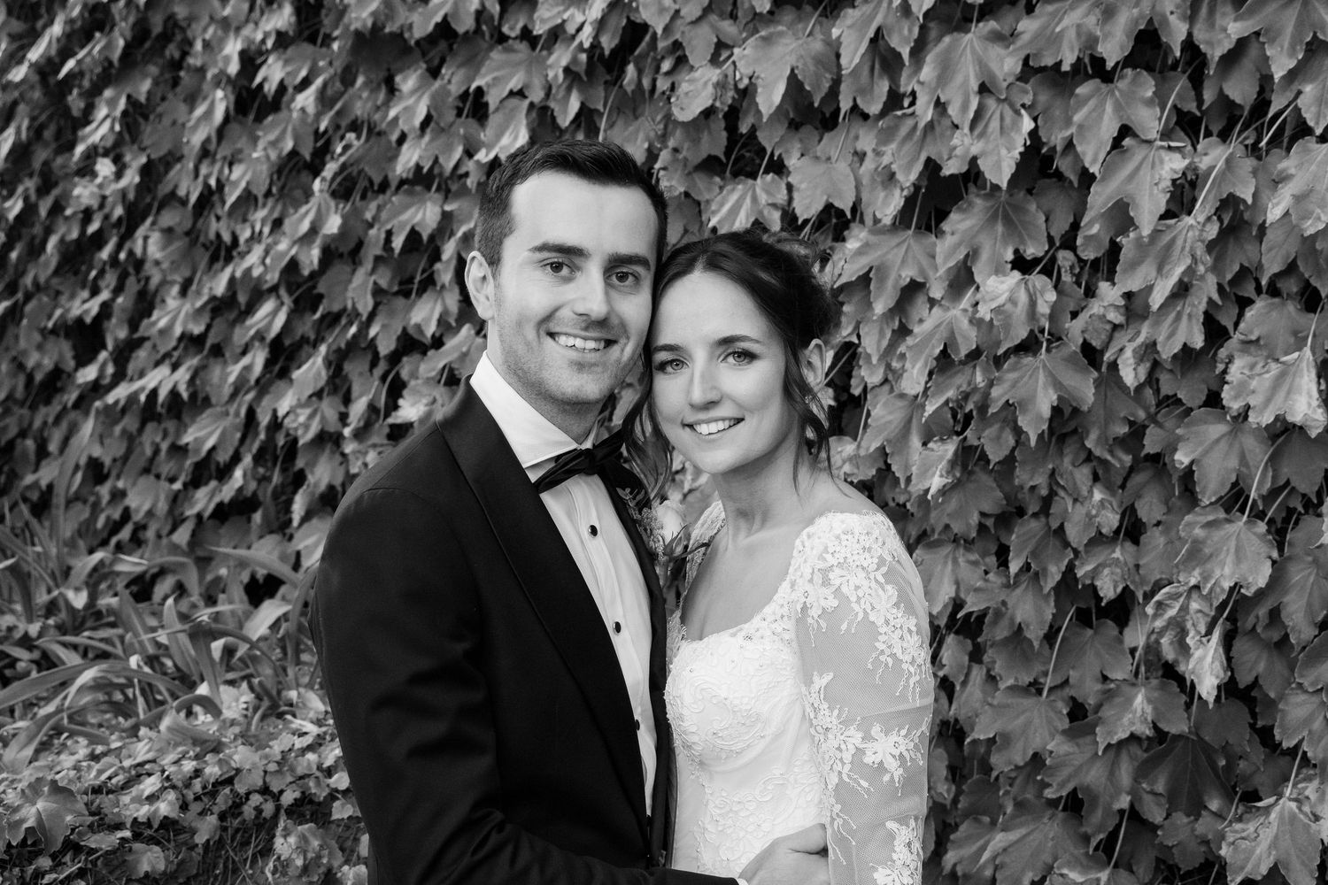 A couple in formal wedding attire poses together against a lush ivy-covered wall backdrop.