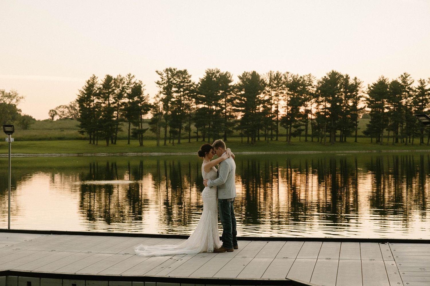 Couple shares an intimate moment on a dock at sunset with a serene lake and tree line in the background.