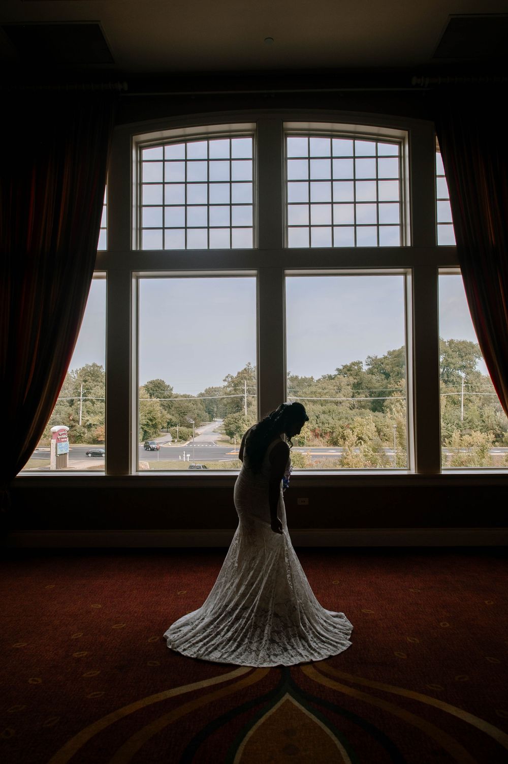 Silhouette of a bride in a flowing wedding dress standing by a large arched window with dramatic lighting.