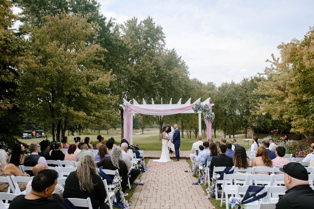 Outdoor wedding ceremony setup with white chairs and guests gathered in a park setting under tall trees.