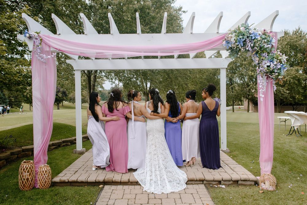 Wedding party gathered under a white pergola decorated with pink fabric and floral arrangements in an outdoor garden setting.