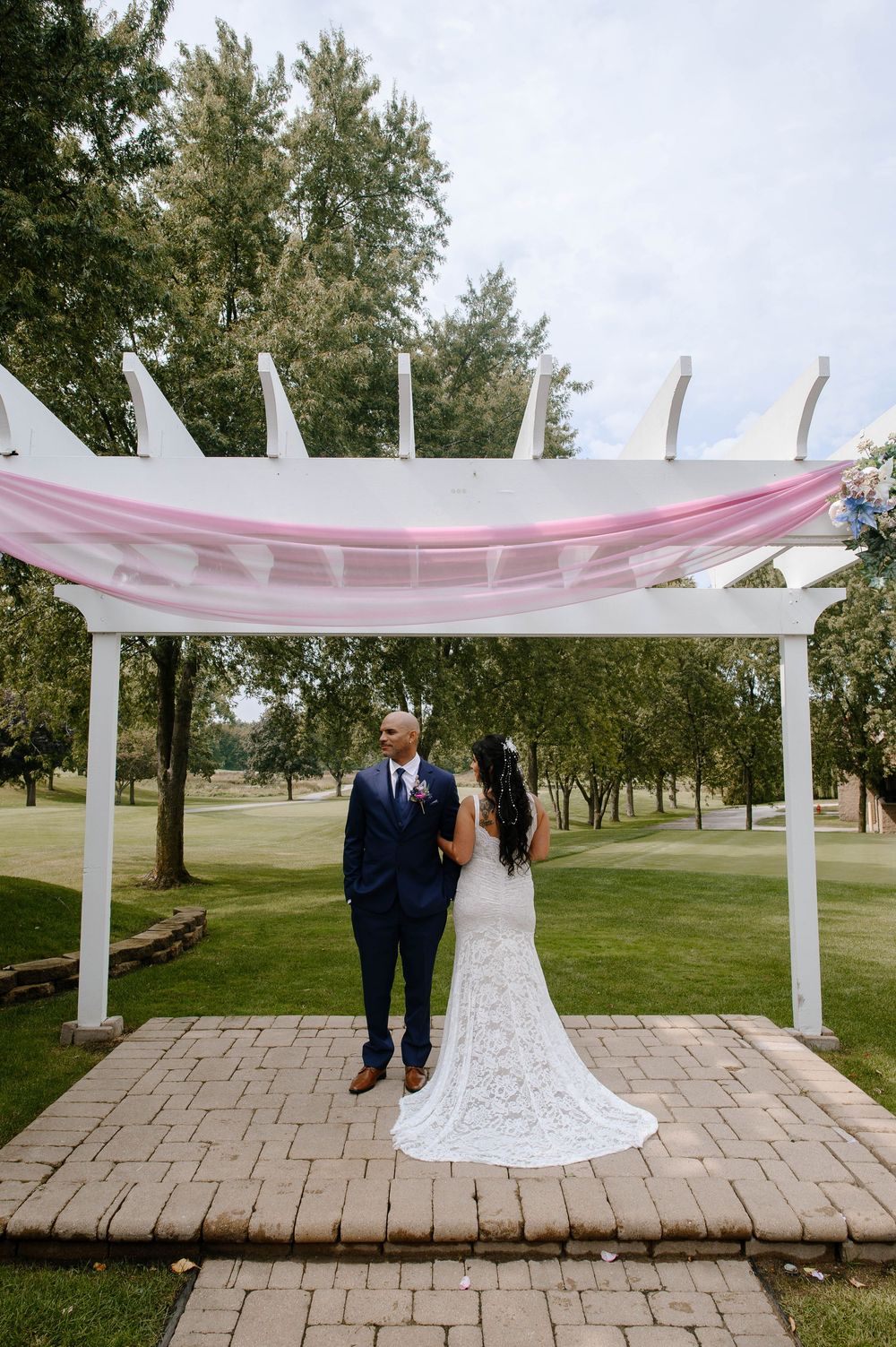 A couple in formal attire stands under a white pergola on a stone patio surrounded by green trees at an outdoor venue.