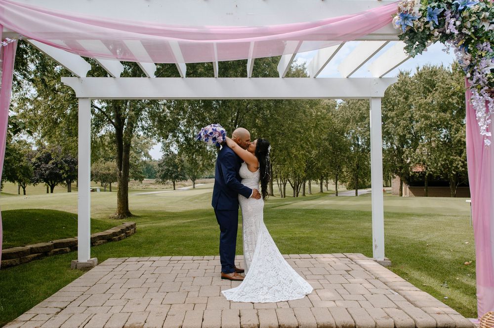A couple embraces under a white pergola decorated with pink fabric draping at an outdoor wedding venue.