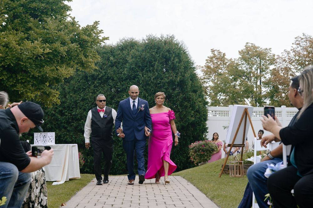 Wedding guests walk down an outdoor stone path during a garden ceremony while photographers capture the moment.