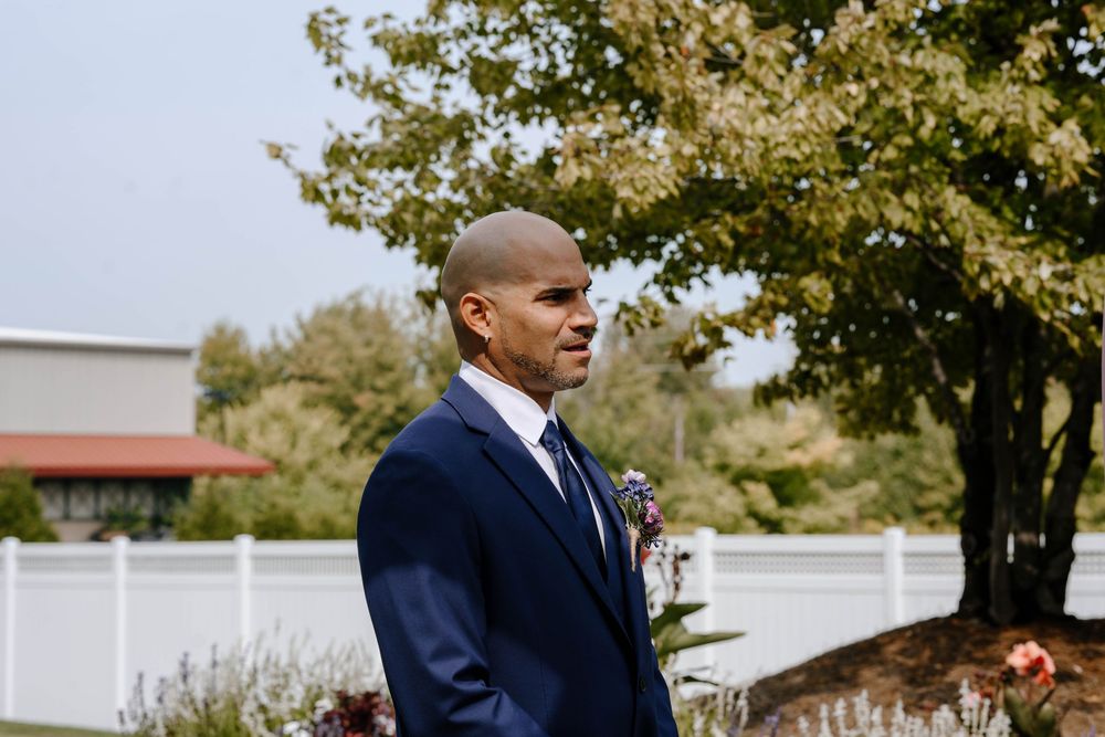 A person in a navy suit walks along a path outdoors with trees and buildings visible in the background.