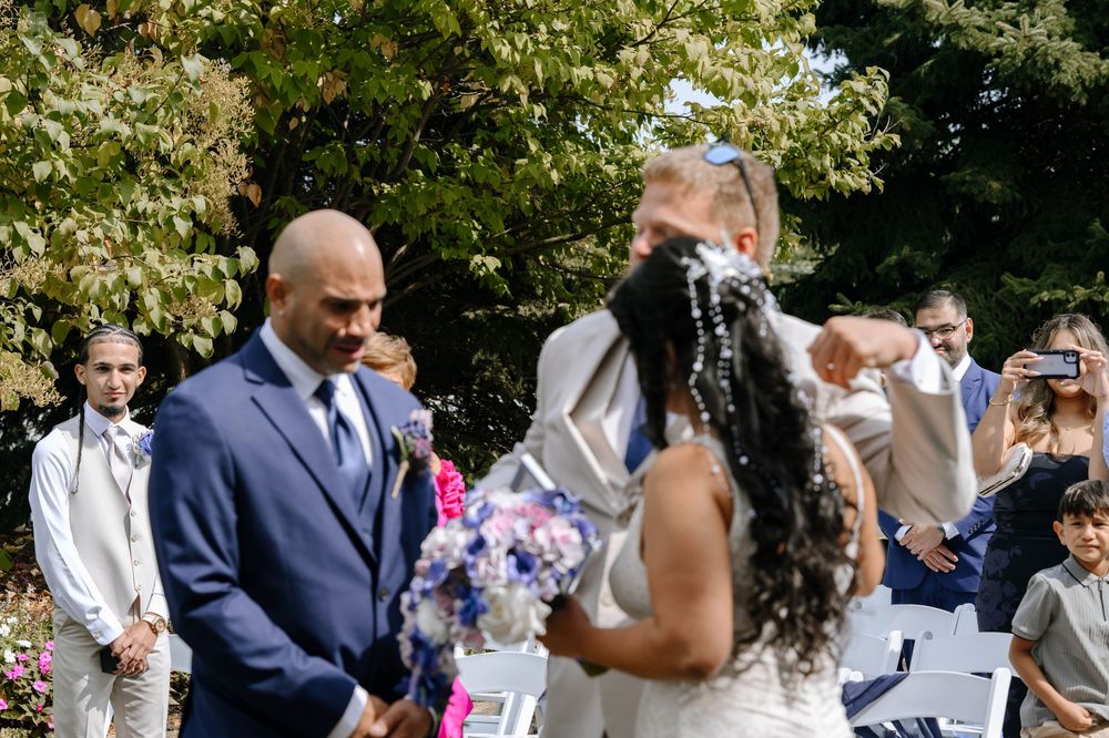 Wedding guests gather outdoors on a sunny day for a garden ceremony with white chairs.