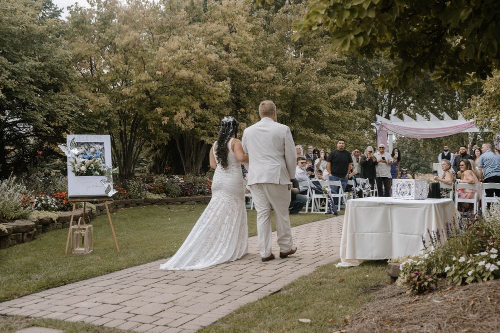 A couple in wedding attire walks down a stone path at an outdoor garden reception with guests seated at tables.