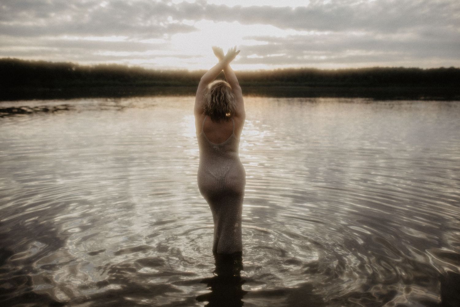 A person stands in a serene lake at sunset with their arms raised in a peaceful gesture.