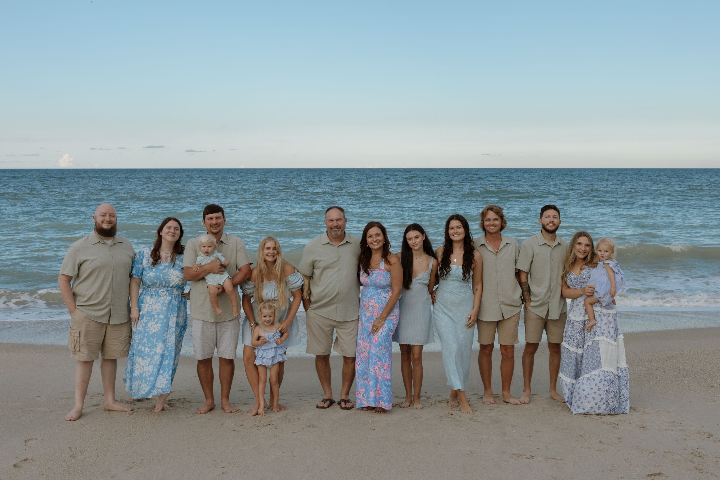 A group in coordinated neutral and blue outfits poses together on a sandy beach at sunset with the ocean behind them.