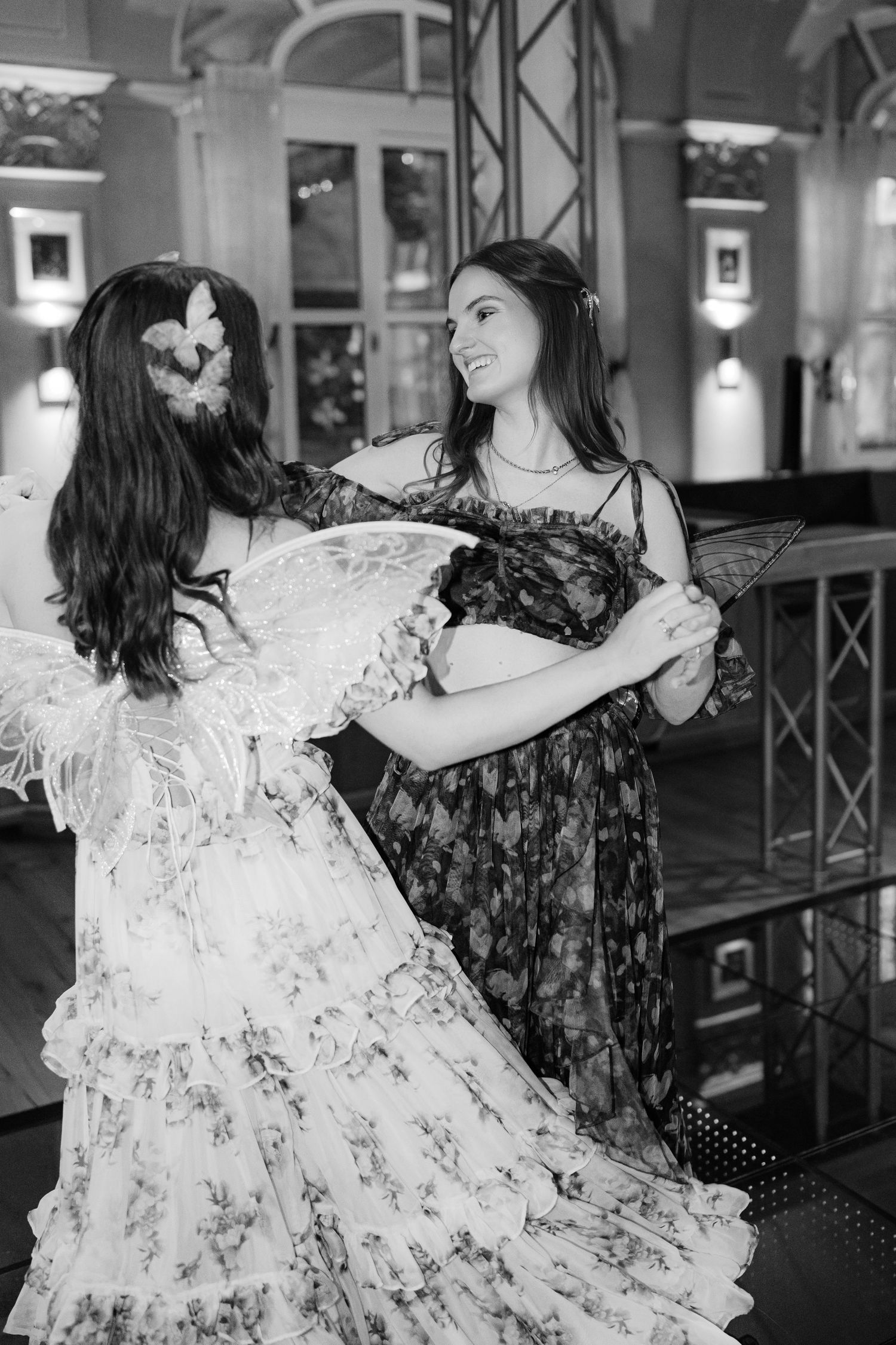 Two people in floral dresses dance together in an elegant indoor venue with warm lighting and wooden accents.
