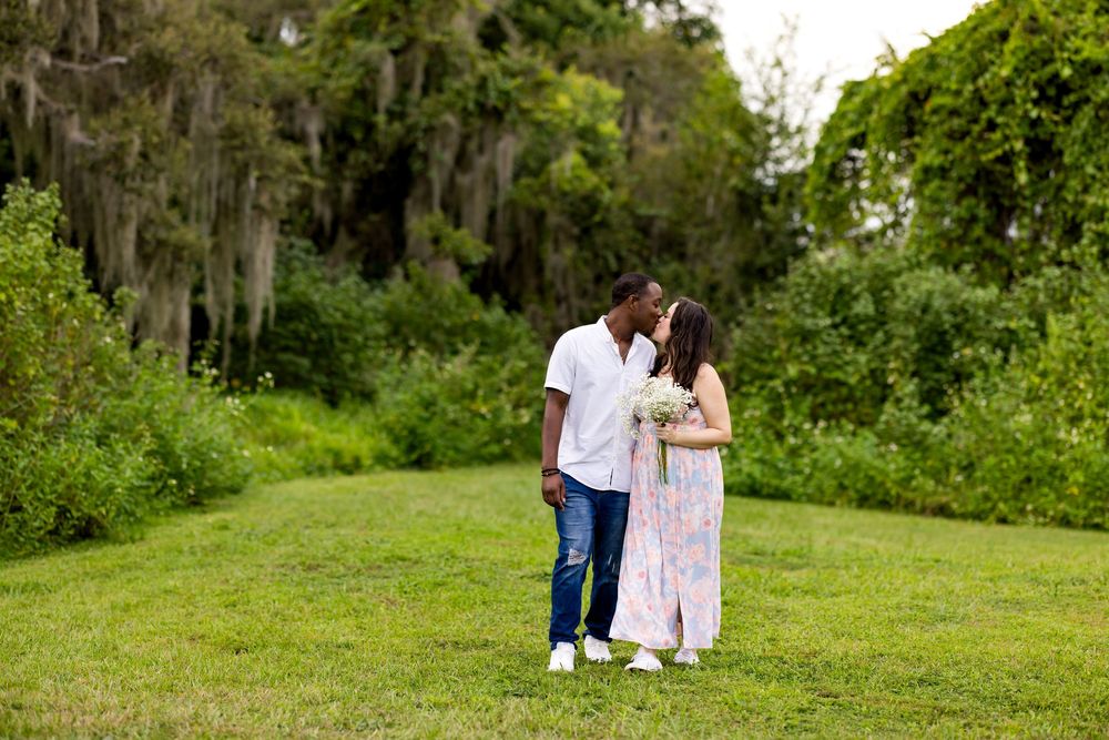 Two people in white and pink outfits sit together on grassy hillside in a beautiful garden setting.