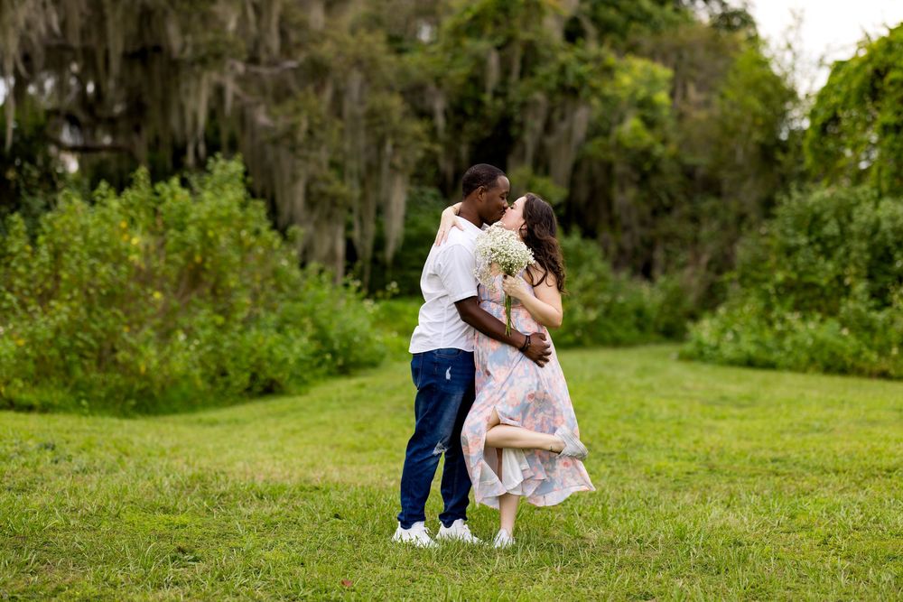 A couple embraces in an intimate moment outdoors in a lush green field surrounded by trees.