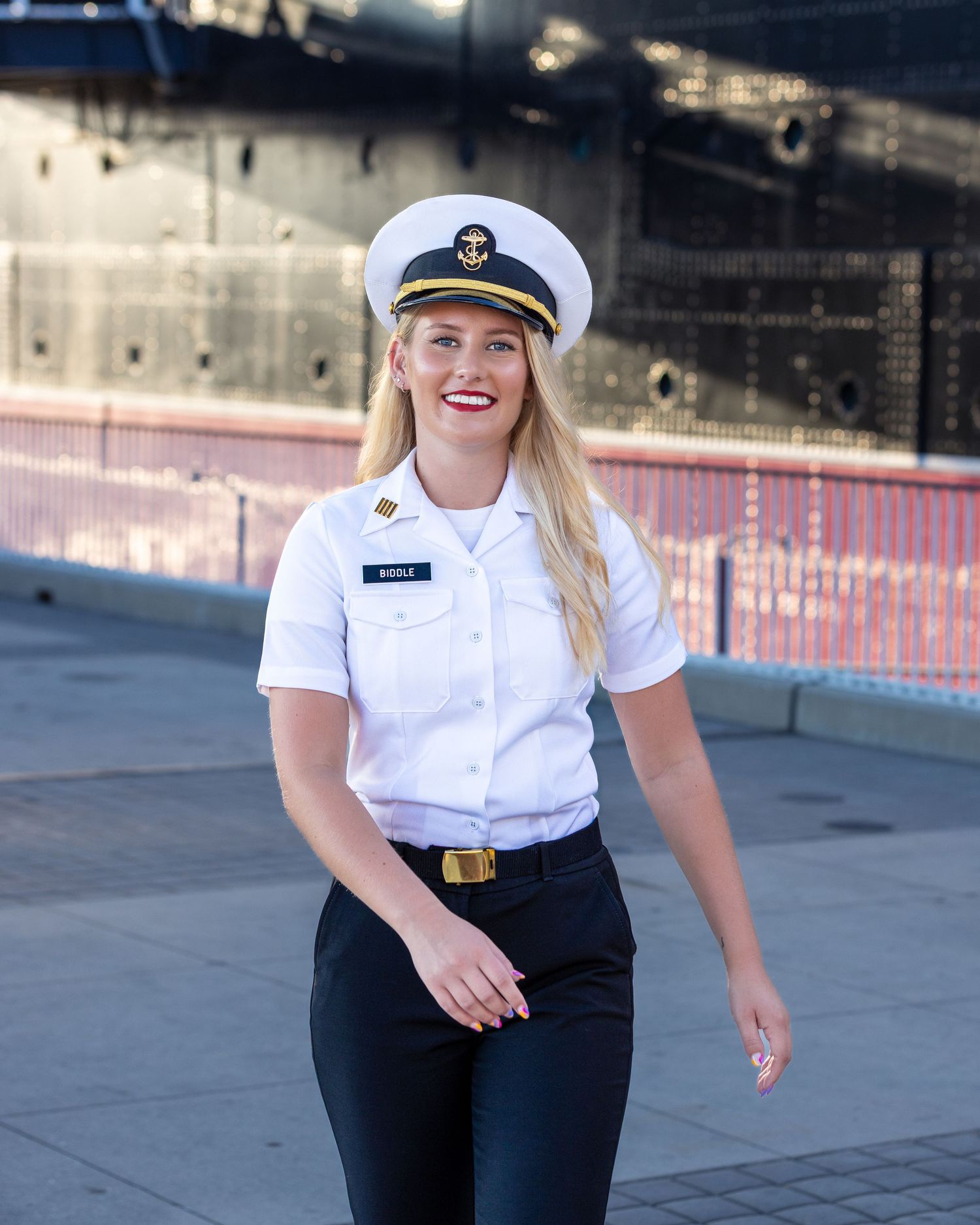 Naval officer in dress whites and dark pants stands outdoors against a blurred background.