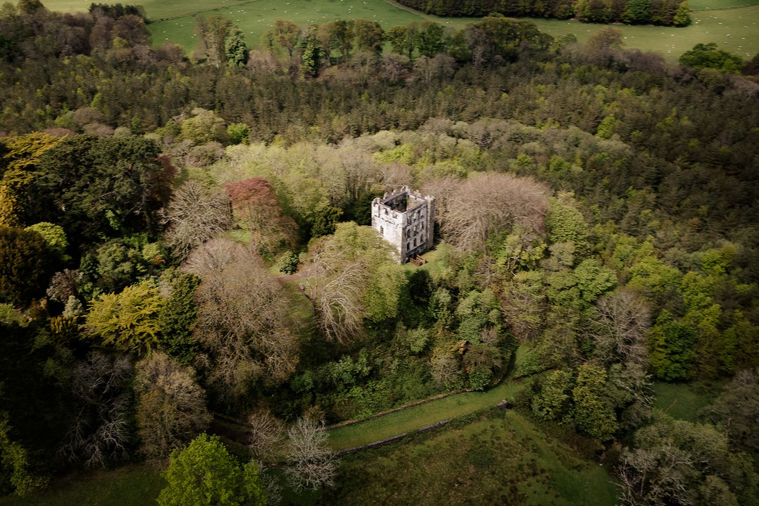 Aerial view of a stone tower or castle hidden among dense forest trees in a rural countryside landscape.