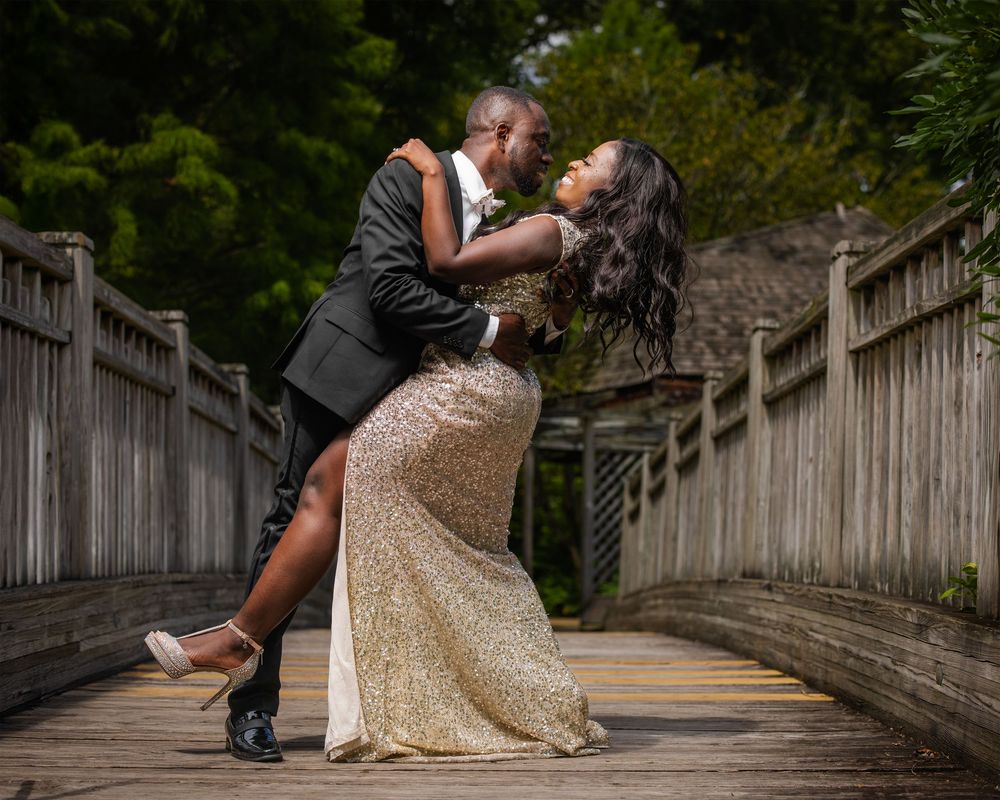 Couple in formal attire posing playfully on wooden bridge in garden setting.