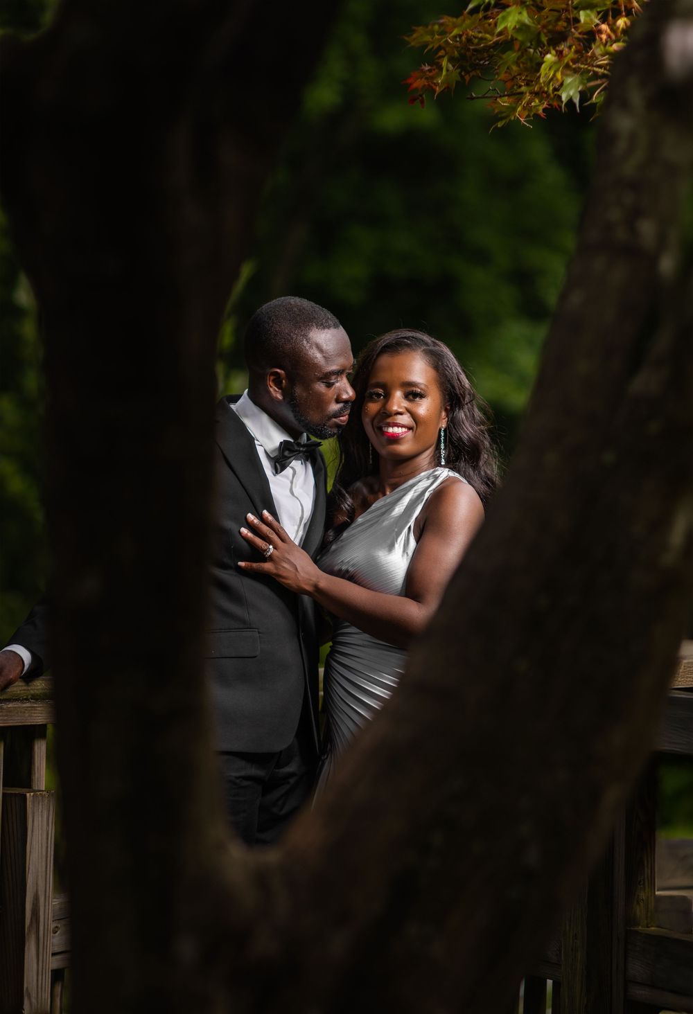 Intimate couple portraits through trees on wooden deck in natural lighting.