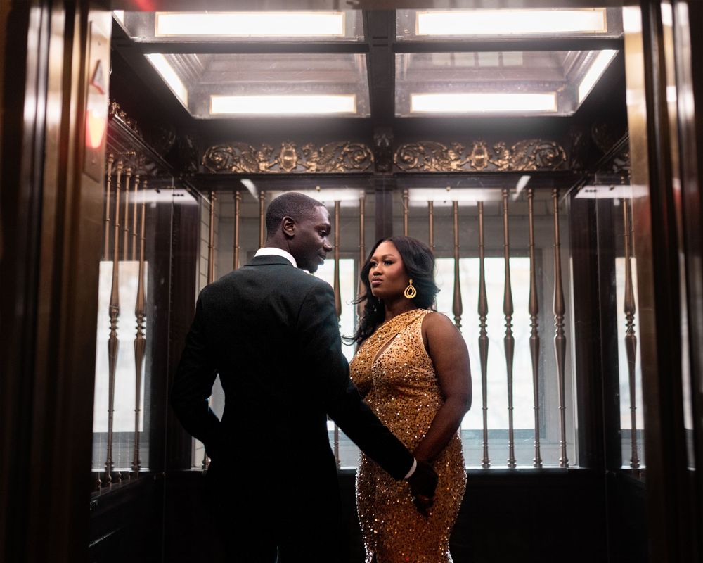 A couple in formal attire poses intimately in an elegant hallway with dramatic lighting and architectural details.