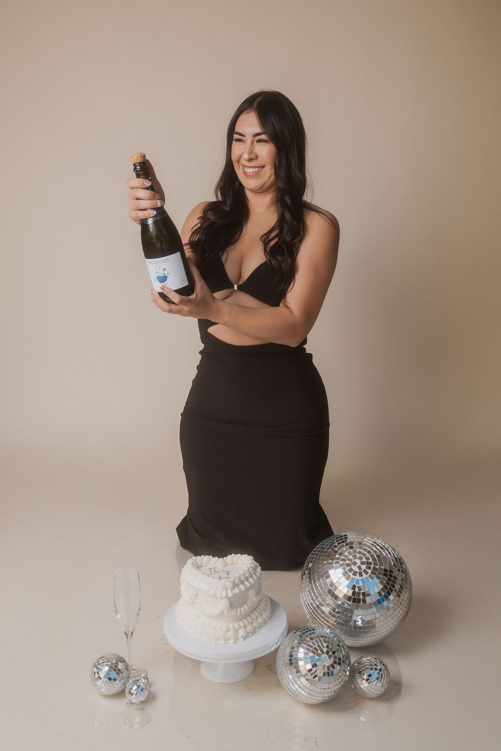 Woman in black dress celebrates with champagne bottle, disco balls and white cake on beige background.