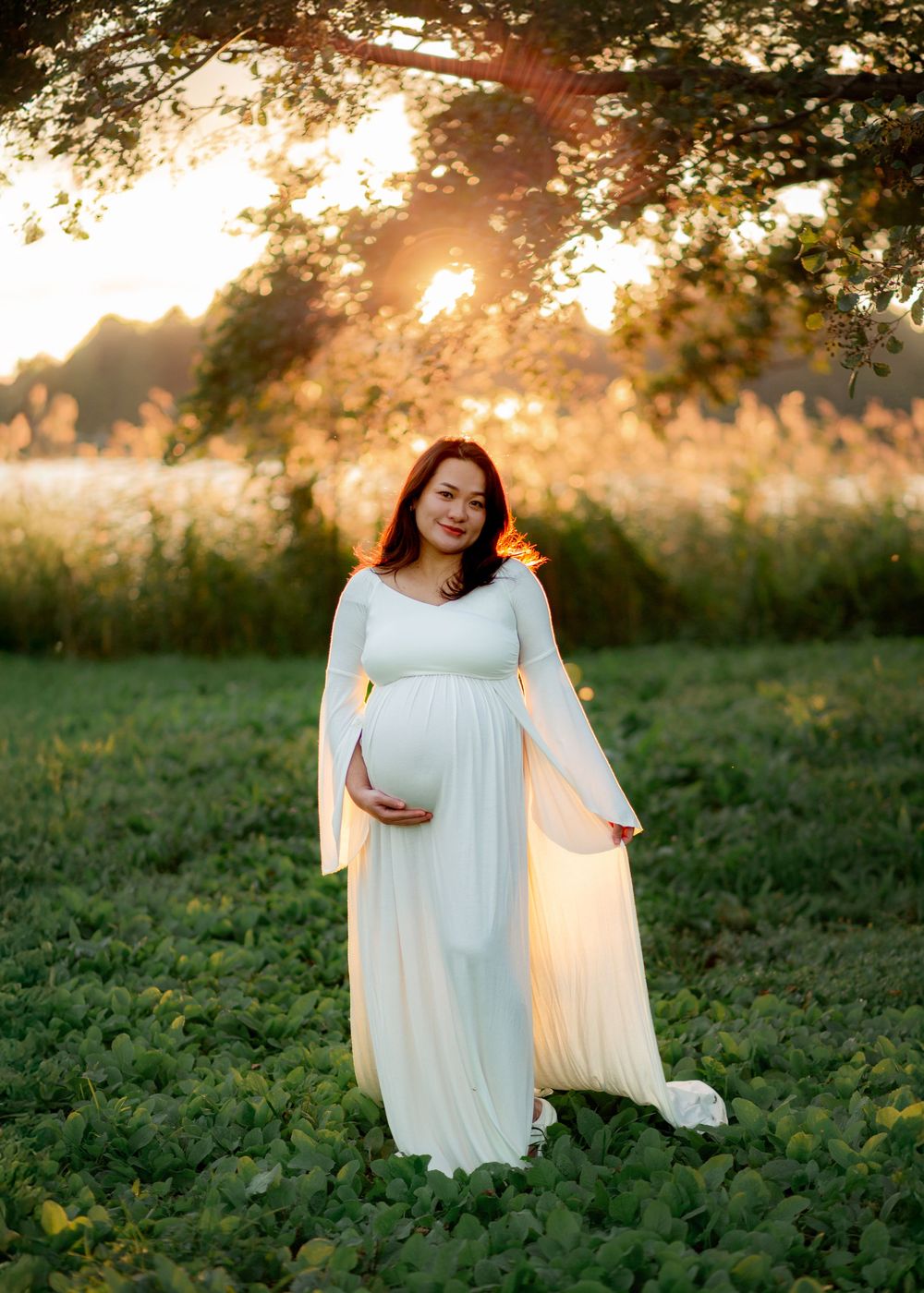 Pregnant woman in white flowing gown cradling her belly in a sunlit green field at golden hour.