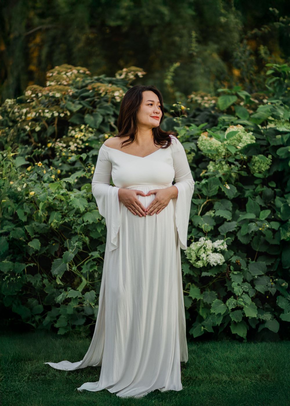 Pregnant woman in off-shoulder white gown making a heart shape over her belly in a lush garden.