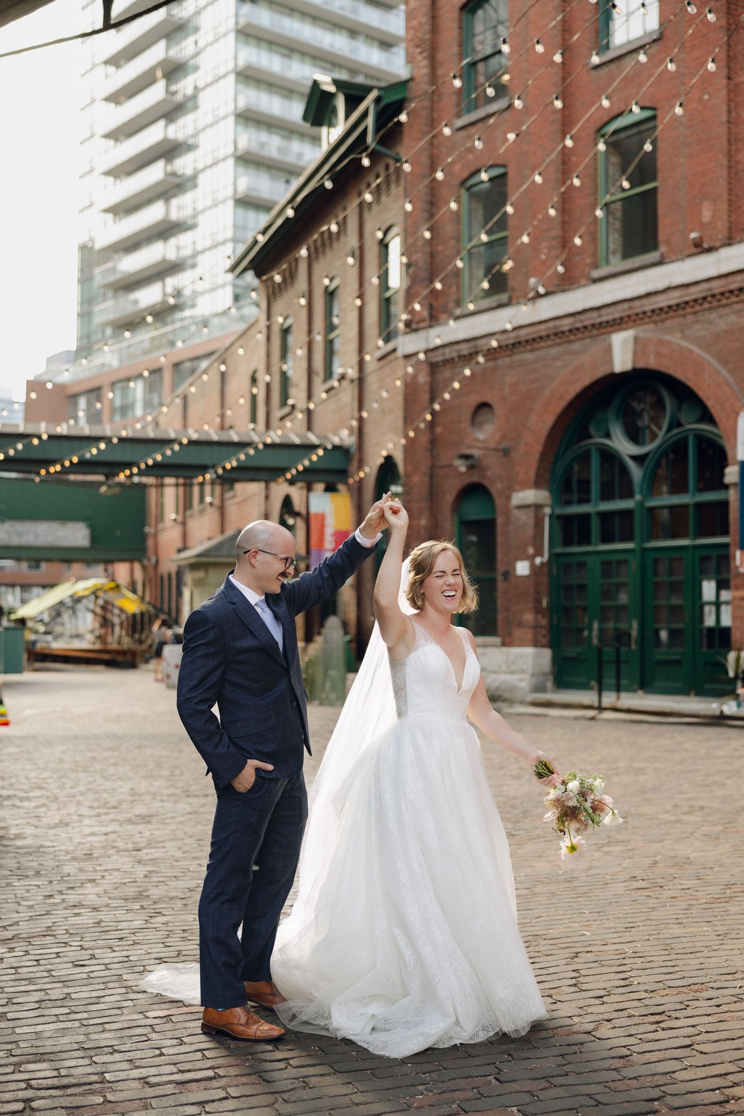 A couple in wedding attire share a romantic moment on cobblestone streets with brick buildings in the background.